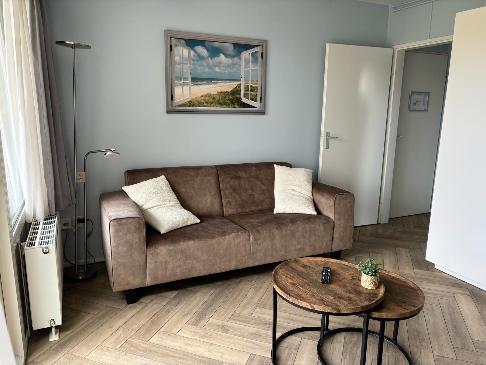 Woonkamer met aan de linkerkant een zwarte bank, in het midden een grijze tafel en aan de rechterkant een tv.