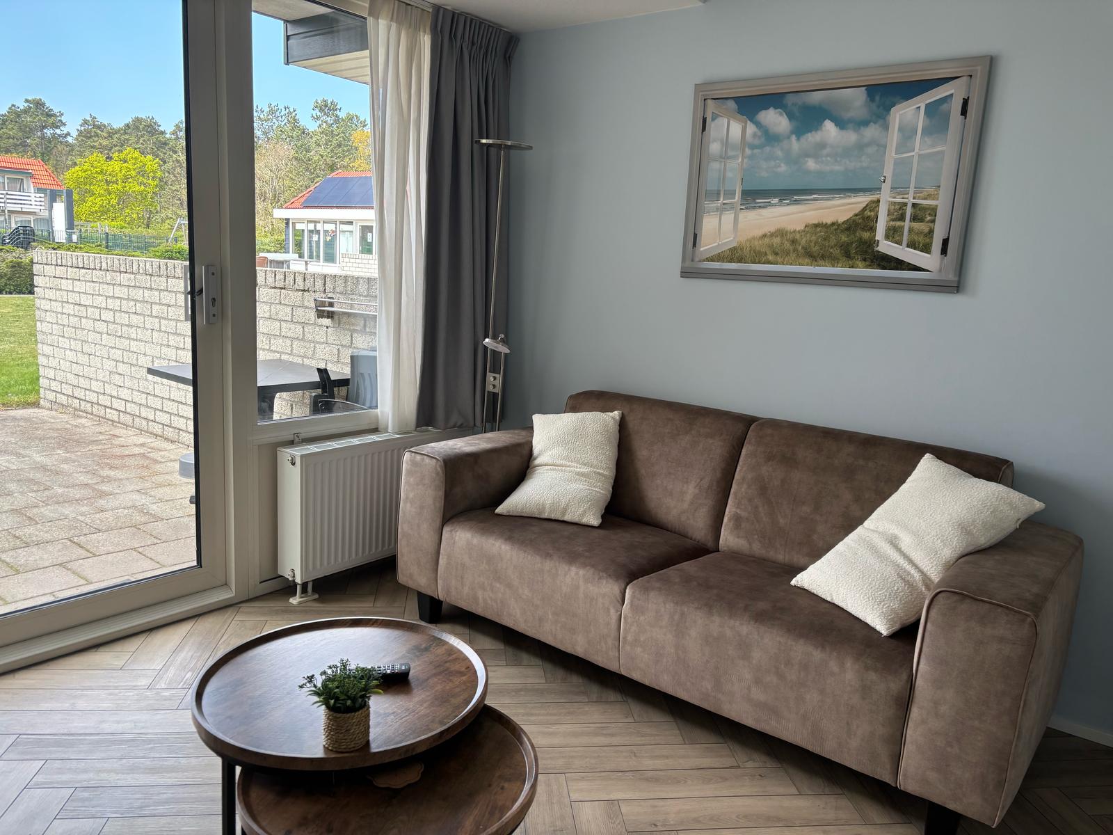 Woonkamer beige bank met schilderij - open raam met uitzicht op zee - uitnodiging om er lekker op uit te gaan.