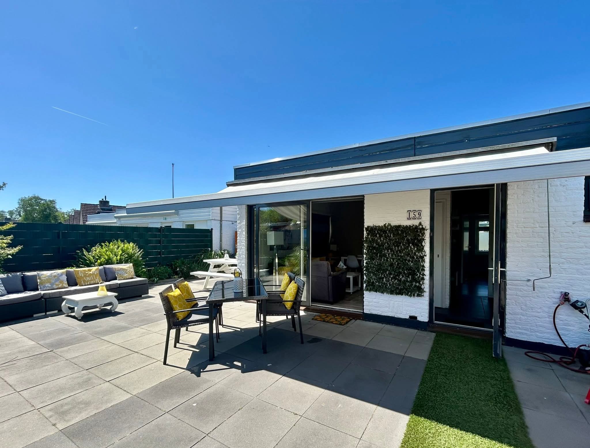 Foto van de bungalow vanaf het straatje; blauwe lucht, met deel van de tuin en terras.