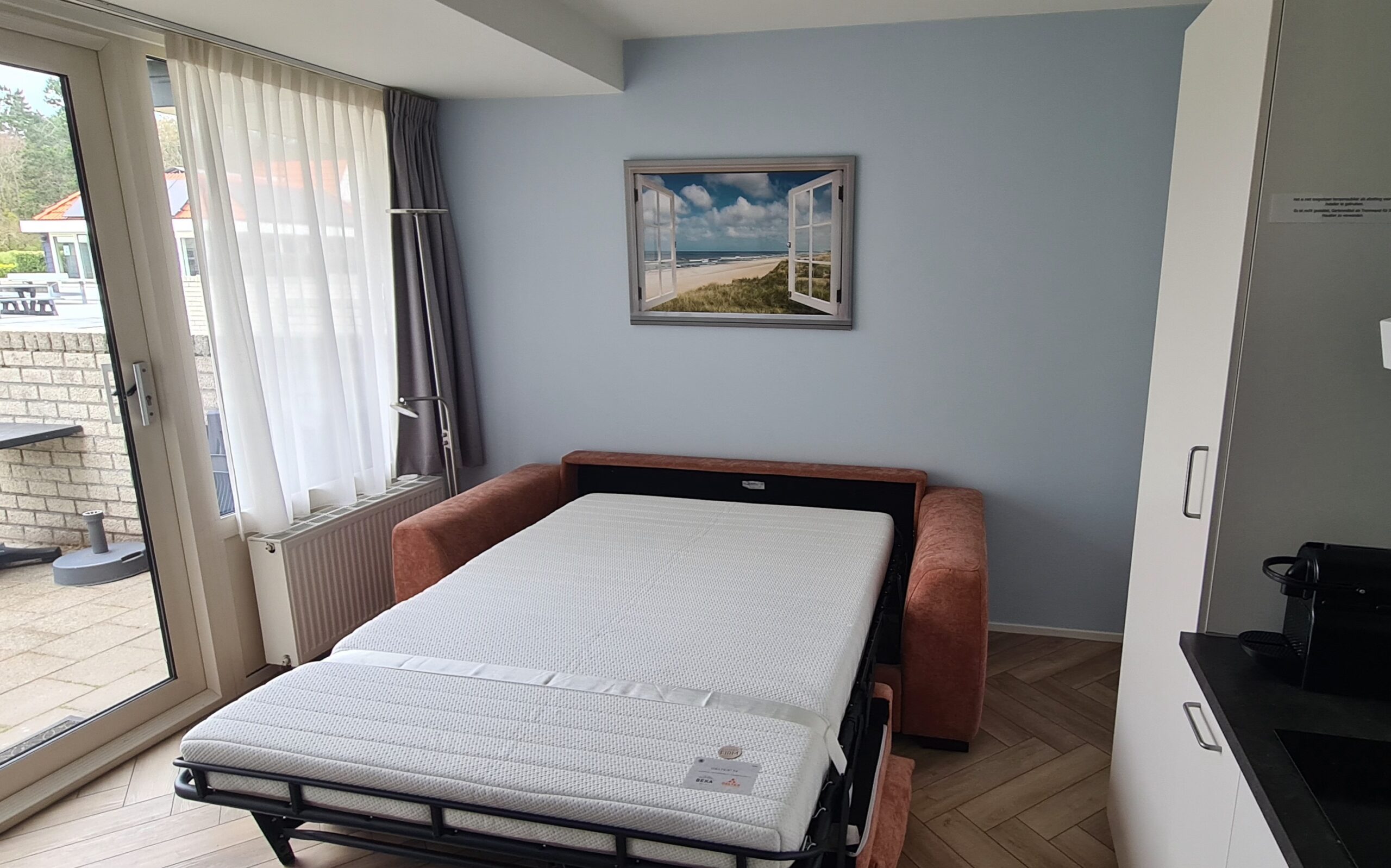 Uitgerolde slaapbank in de woonkamer met schilderij - open raam met uitzicht op zee - op de achtegrond.